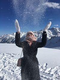 Young woman throwing snow while standing on snowcapped mountain against blue sky