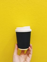 Cropped hand holding disposable cup against yellow wall