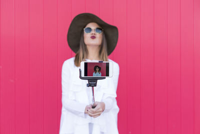 Young woman taking selfie while standing against pink wall