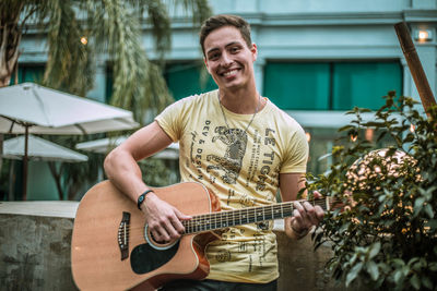 Portrait of young man playing guitar while standing against building