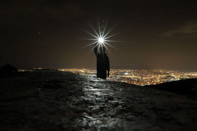 Silhouette person standing on illuminated light against sky at night