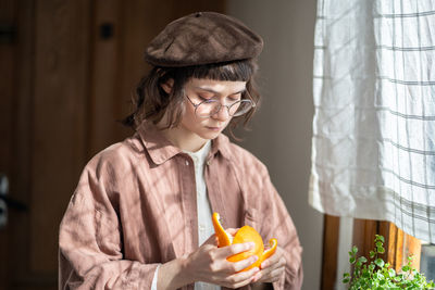 Focused teen girl in vintage style clothes, beret, eyeglasses peeling orange at home near window.