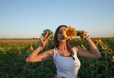 Portrait of young woman holding sunflower gesturing against sky during sunset