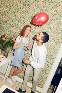 Tilt shot of young man playing with balloon while standing by woman against wallpaper during dinner party at home