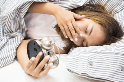 Woman yawning while holding alarm clock on bed