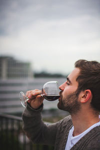 Side view of young man drinking red wine against sky
