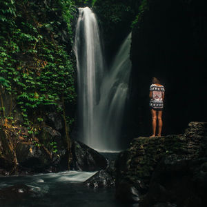 Rear view of woman standing on rock by waterfall