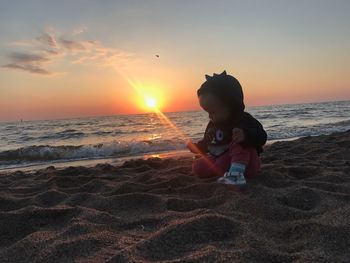Cute girl sitting at beach against sky during sunset