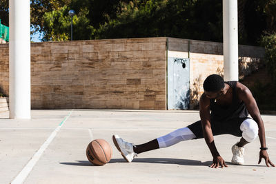 Fit african american male basketball player warming up legs and doing side lunges while preparing for training on sports grond in summer