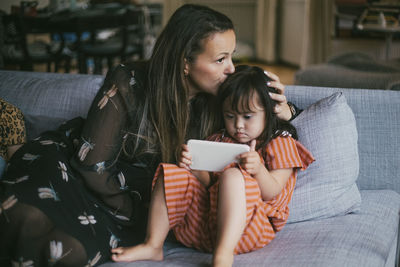 Mother kissing down syndrome daughter using smart phone on sofa at home