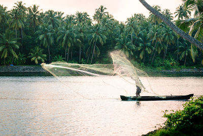 Fisherman holding fishing net in boat on lake against palm tree