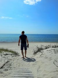 Rear view of man standing at beach against blue sky during sunny day