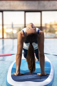 Female yoga instructor in bridge position on paddleboard over swimming pool