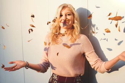 Portrait of smiling young woman standing amidst leaves against wall