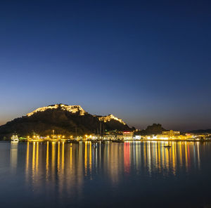 Illuminated buildings by sea against clear sky