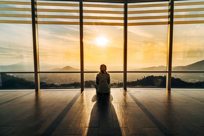 Rear view of young woman looking through window during sunset