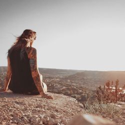 Rear view of woman looking at landscape while sitting on rock against clear sky