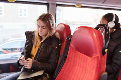 Teenage girl using smart phone and female listening music through headphones while traveling in bus