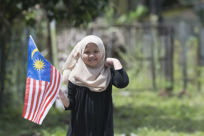 Portrait of girl in hijab holding malaysian flag while standing at field
