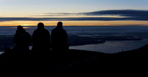 Silhouette people sitting by sea against sky during sunset