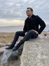 Full length of man sitting on rock by sea against sky