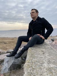 Young man sitting on rock by sea against sky