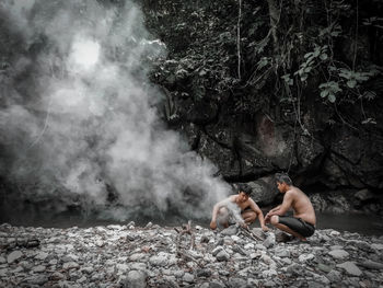 Shirtless male friends crouching by campfire at riverbank