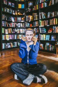 Young woman hiding face with book while sitting cross-legged in bookstore
