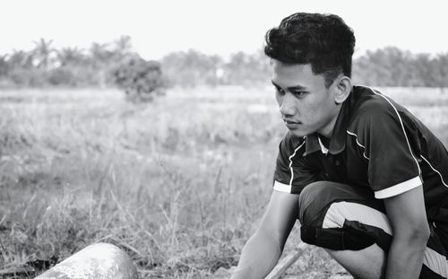 Young man looking away while crouching on grassy field against sky