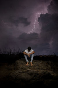 Man sitting on rock against storm clouds at night