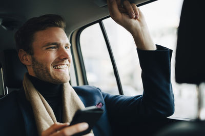 Smiling businessman with phone looking away while sitting in taxi