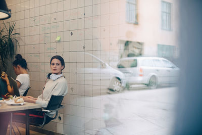 Young businesswoman looking away while sitting at desk in small office seen through window