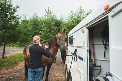 Senior man touching horse tied to horse trailer on road