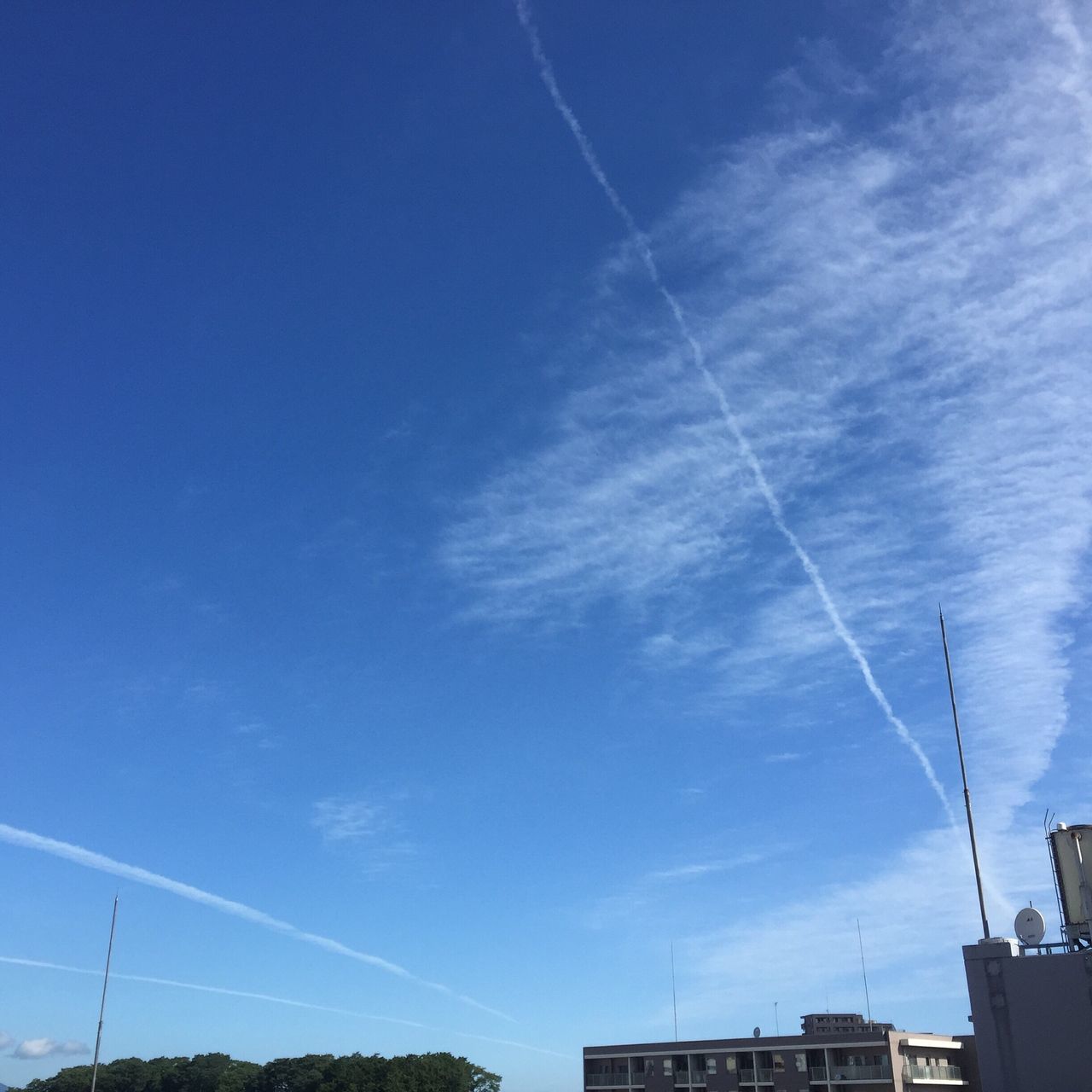 vapor trail, low angle view, blue, architecture, sky, building exterior, built structure, city, high section, cable, cloud - sky, cloud, day, outdoors, power cable, scenics, power line, city life, no people, apartment, long, beauty in nature, wispy, town, tranquility