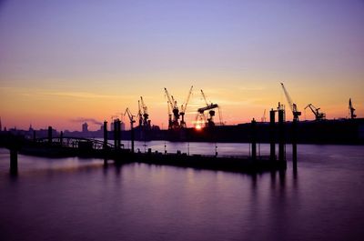 Silhouette cranes at harbor against sky during sunset