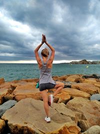 Rear view of woman doing yoga while standing on rock at beach against sky