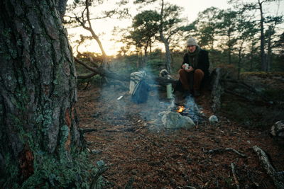 Full length of man sitting on fallen tree by campfire at forest