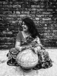 Smiling woman in sari playing music on pot while sitting against wall