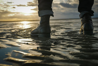 Low section of man standing at beach against sky during sunset