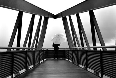 Rear view of young man with umbrella standing on bridge against cloudy sky