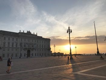 People walking on street by buildings against sky during sunset