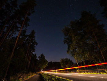 Light trails on road amidst trees against sky at night