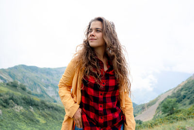 A young beautiful woman with long curly hair stands against the backdrop of a mountain landscape.