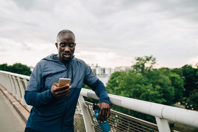 Sportsman using mobile phone while leaning on railing at footbridge against sky