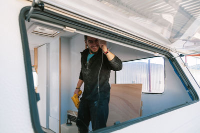 Portrait of smiling man holding drill seen through window of camper trailer