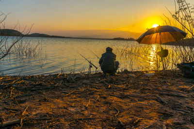 Rear view of man sitting on beach against sky during sunset