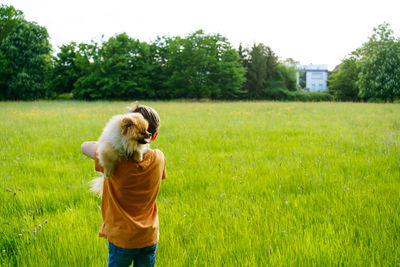 A boy runs through a summer field with small pomeranian dog with new house in background