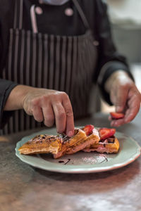 Crop anonymous male cook putting ripe strawberry slices on viennese waffles with chocolate sauce and powdered sugar on plate in kitchen