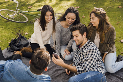 Smiling friends using mobile phone while enjoying picnic at park