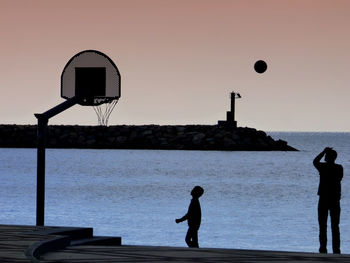 Silhouette of basketball players by sea against sky during sunset